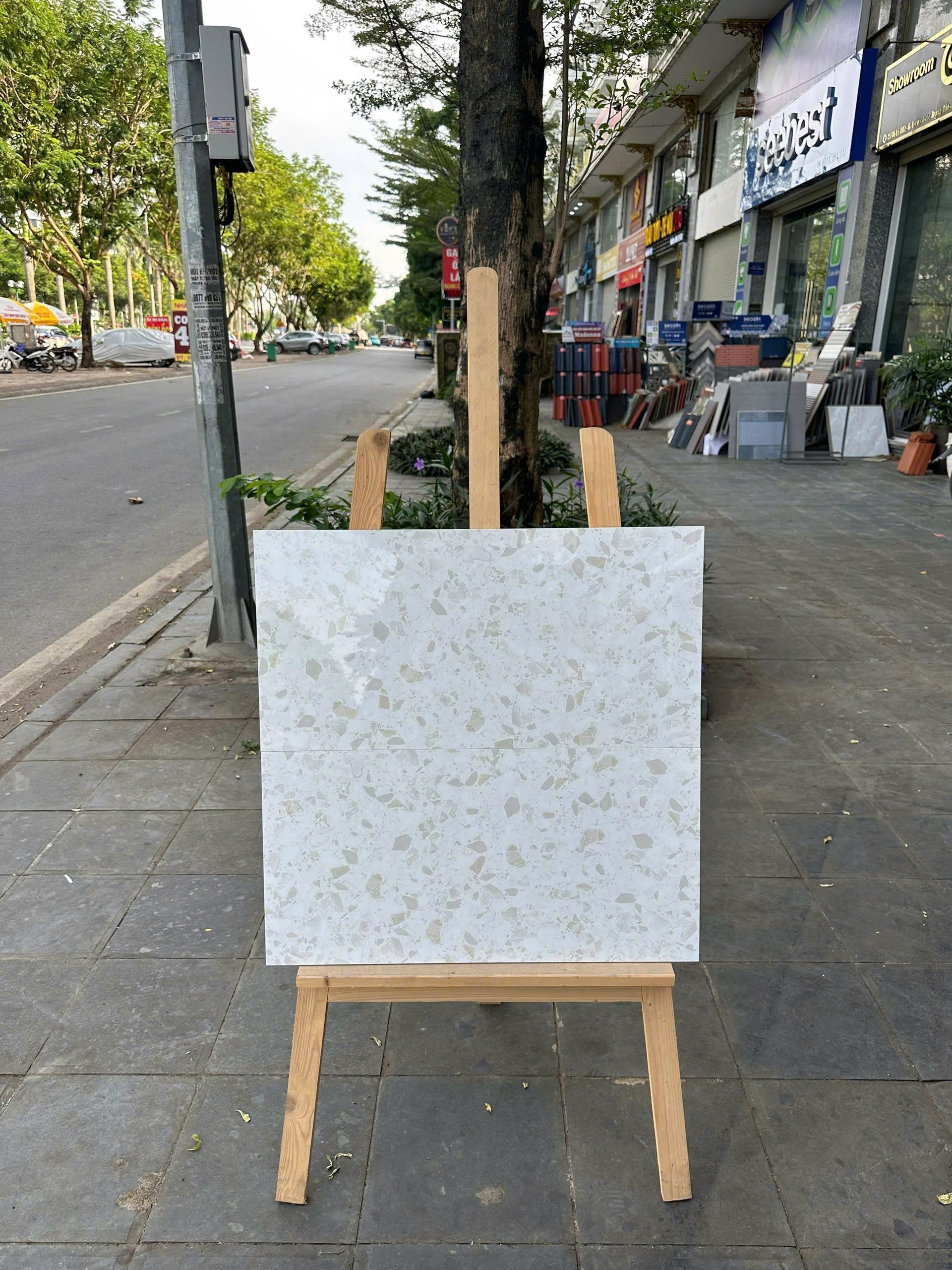 Tấm gạch trắng với hoa văn xám nhạt đặt trên giá gỗ ngoài vỉa hè, nền là đường phố và cây xanh.